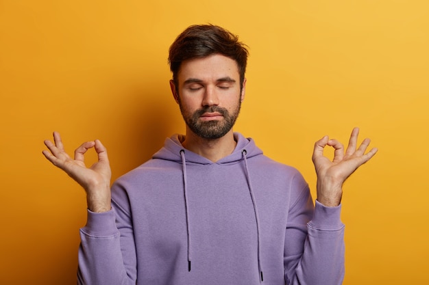 Peaceful patient bearded man raises hands sideways with zen gesture, keeps eyes closed, rests after work or studying, being patient, poses against yellow wall, breathes deeply and feels relieved