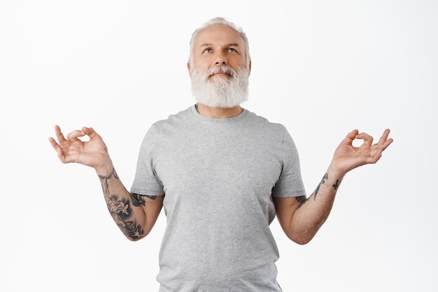 Мирный пожилой мужчина чувствует дзен, медитирует и отдыхает, дышит спокойно и смотрит вверх, улыбаясь, удовлетворенно успокаиваясь, держась за руки в знаках мудры йоги, стоящих на белом фоне