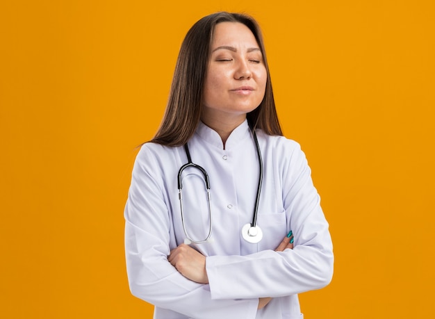 Peaceful young asian female doctor wearing medical robe and stethoscope standing with closed posture and closed eyes isolated on orange wall with copy space