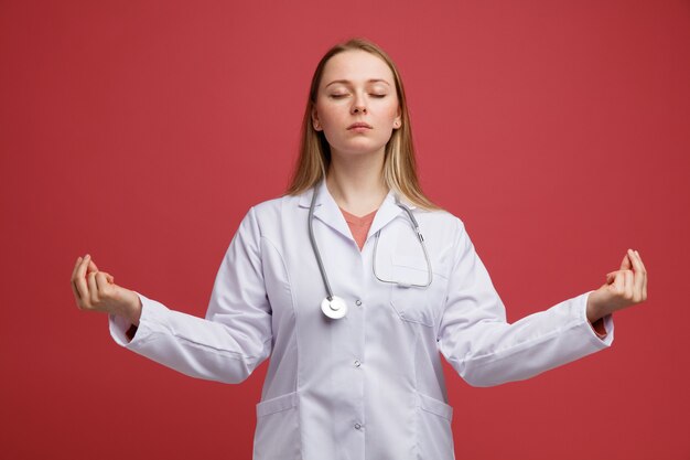 Peaceful young blonde female doctor wearing medical robe and stethoscope around neck meditating with closed eyes 