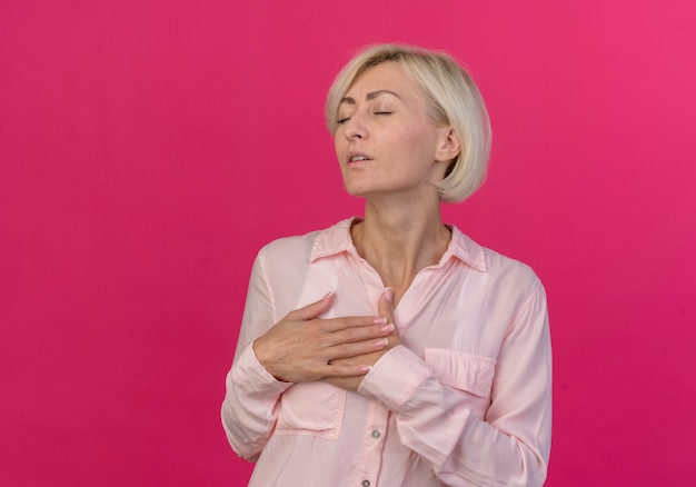 Free photo peaceful young blonde slavic woman keeping hands on chest with closed eyes isolated on pink background with copy space