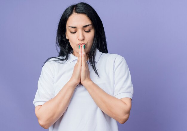 Peaceful young brunette caucasian girl holds hands together with closed eyes isolated on purple wall