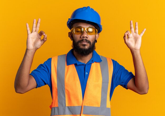 peaceful young builder man in safety glasses wearing uniform with safety helmet pretending to meditate isolated on orange wall with copy space