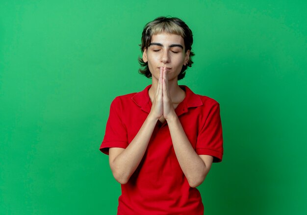 Peaceful young caucasian girl with pixie haircut putting hands in pray gesture praying with closed eyes isolated on green background with copy space