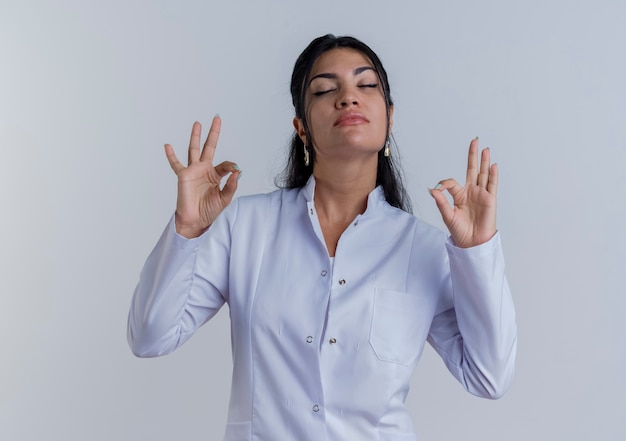 Peaceful young female doctor wearing medical robe doing ok sings with closed eyes isolated on white wall