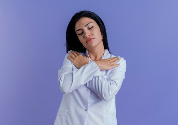 의료 가운을 입고 평화로운 젊은 여성 의사가 복사 공간이 보라색 벽에 고립 된 닫힌 눈으로 어깨에 넘어 손을 유지
