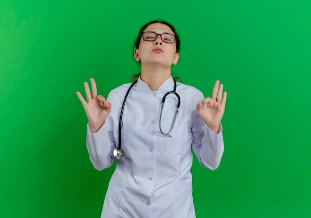 의료 가운과 청진 기 및 복사 공간 녹색 벽에 고립 된 닫힌 눈으로 확인 서명을 하 고 안경을 착용하는 평화로운 젊은 여성 의사