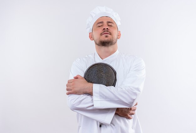 Peaceful young handsome cook in chef uniform holding frying pan with closed eyes on isolated white space