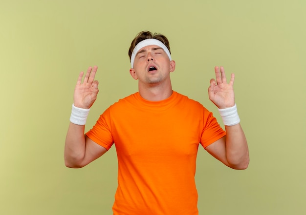 Peaceful young handsome sporty man wearing headband and wristbands doing ok signs with closed eyes isolated on olive green background