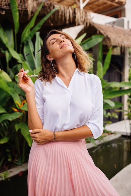 Peaceful young happy woman with short curly hair in pink long skirt and white shirt alone outside her villa