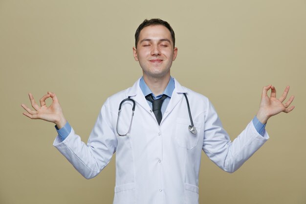 Peaceful young male doctor wearing medical robe and stethoscope around neck meditating with closed eyes isolated on olive green background