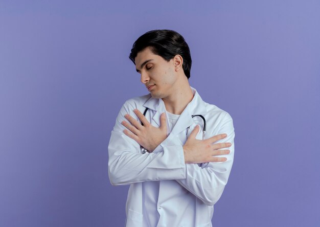 Peaceful young male doctor wearing medical robe and stethoscope keeping hands crossed on arms turning head to side with closed eyes isolated