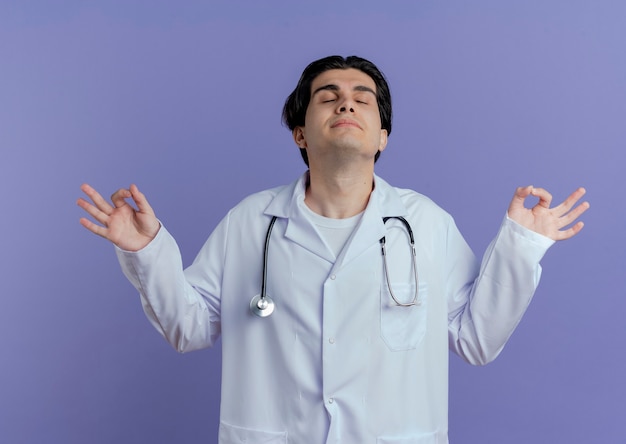 Peaceful young male doctor wearing medical robe and stethoscope meditating with closed eyes isolated on purple wall