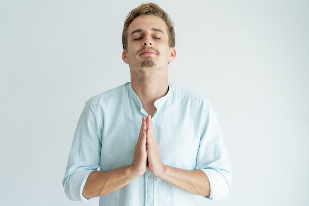 Peaceful young man praying with his eyes closed and keeping hands together.