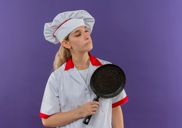 Мирный молодой симпатичный повар в униформе шеф-повара держит сковороду с закрытыми глазами, изолированную на фиолетовом пространстве