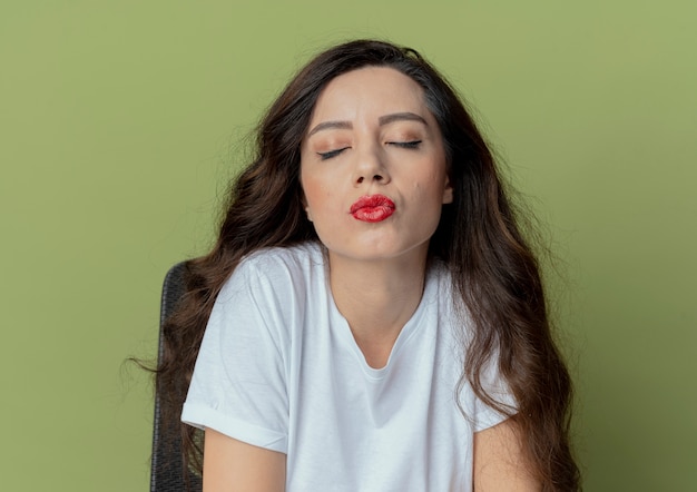 Tranquilla giovane bella ragazza seduta al tavolo per il trucco con strumenti di trucco facendo gesto di bacio con gli occhi chiusi, isolato su sfondo verde oliva