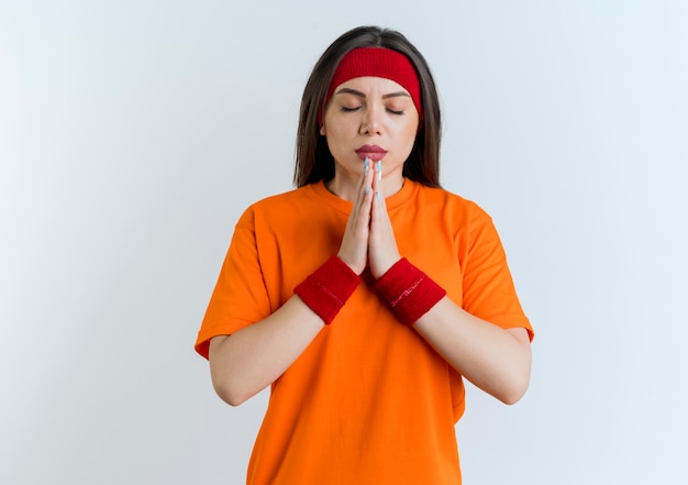 Peaceful young sporty woman wearing headband and wristbands putting hands together praying with closed eyes isolated on white wall with copy space