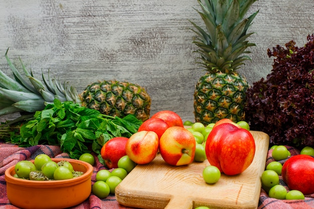 Peaches in a cutting board and clay bowl with green leaves, two pineapples and lettuce side view on picnic cloth and grunge wall