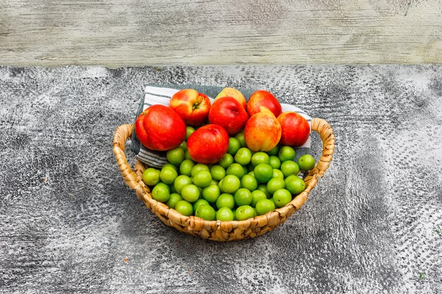 Peaches and greengages in a wicker basket and picnic cloth on grunge grey and wood, high angle view.