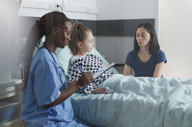 Pediatric clinic medical nurse taking notes about successful treatment scheme. Young considerate kind mother being attentive to nurse speaking about little girl health hazards while in recovery ward.