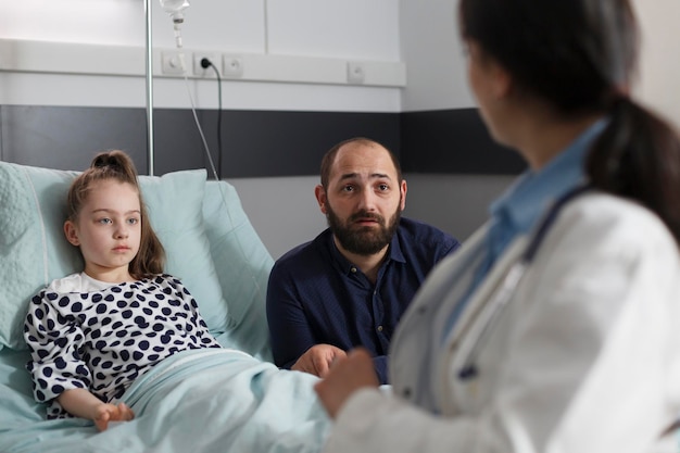 小児医療施設の病棟で休んでいる入院中の病気の少女の父親と話している小児科の専門家。一般開業医は、患者のベッドに座っている体調不良の回復中の子供に相談します。