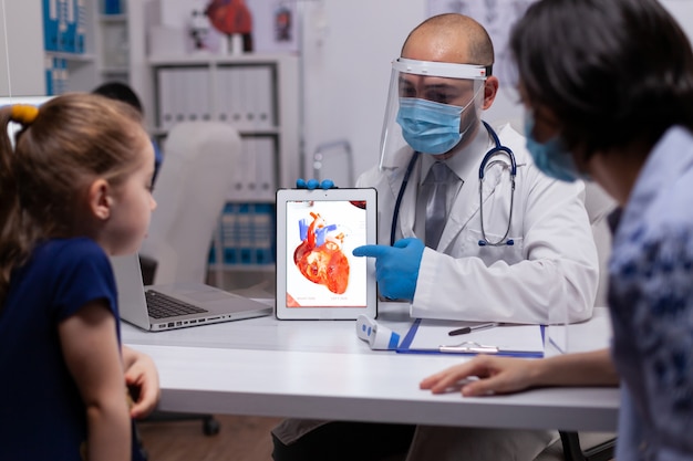 新しい​通常​の​診療所​で​心臓​の​状態​を​説明する​デジタル​タブレット​pc​を​保持している​小児科医​の​医師