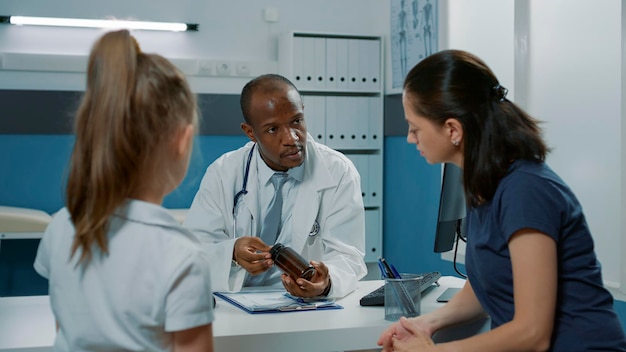 Pediatrician giving bottle of pills to parent of small kid with illness after checkup examination. Medic giving prescription medicine in flask to woman and child, helping with recovery.