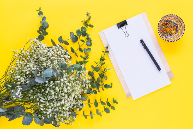 Free Photo pen on clipboard; colorful bowl and baby's breath flowers on yellow backdrop