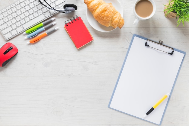 Pen on clipboard with breakfast and office stationeries on white table