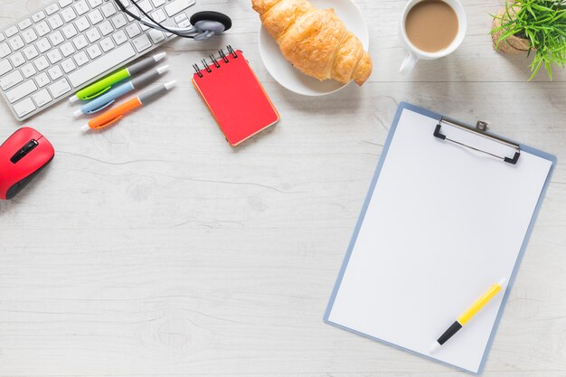 Pen on clipboard with breakfast and office stationeries on white table