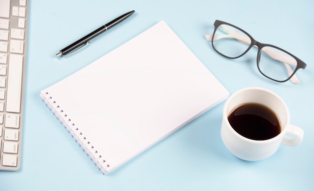 Pen; eyeglasses; coffee cup; keyboard and blank spiral notepad on blue background