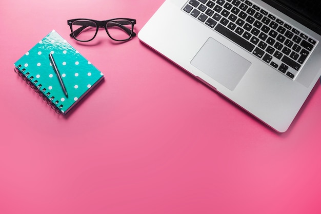 Pen on spiral notebook; eyeglasses and laptop on pink background