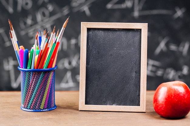 Pen stand and blank chalkboard with apple placed on desk