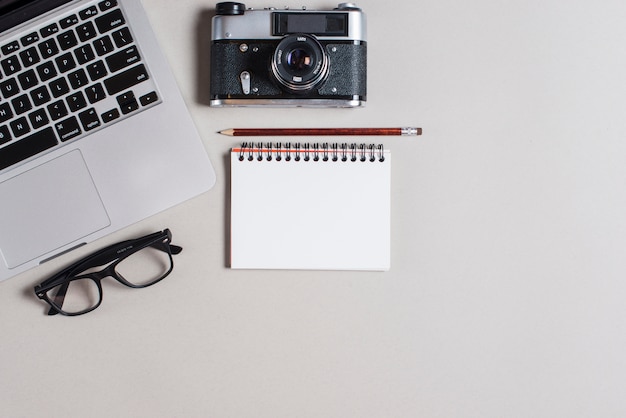 Pencil over spiral notepad with laptop; camera and eyeglasses on gray backdrop