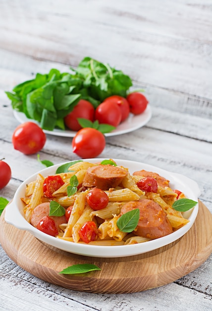 Penne al sugo di pomodoro con salsiccia, pomodori, basilico verde decorato in padella su un tavolo di legno