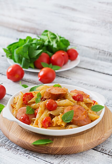 Penne pasta with tomato sauce with sausage, tomatoes, green basil decorated in a frying pan on a wooden table