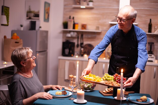 Pensioner couple smiling at each other in kitchen during relationship celebration. Elderly old couple talking, sitting at the table in kitchen, enjoying the meal,