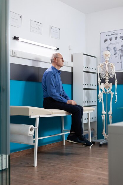 Pensioner senior old man standing on bed waiting for healthcare treatment recovering during medical appointment in hospital office. Medicine services support. Recovery assistance