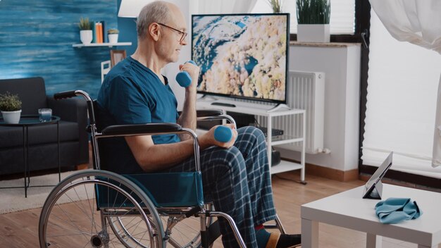 Пенсионер сидит в инвалидной коляске и делает упражнения с гантелями, просматривая видео урока на планшете. Человек с ограниченными физическими возможностями смотрит видео тренировки, поднимая тяжести