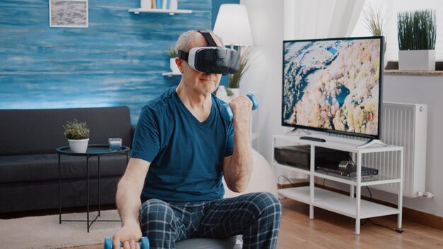 Pensioner using vr glasses to do physical exercise with dumbbells while sitting on fitness toning ball. Senior person with virtual reality goggles training arms muscles with weights.