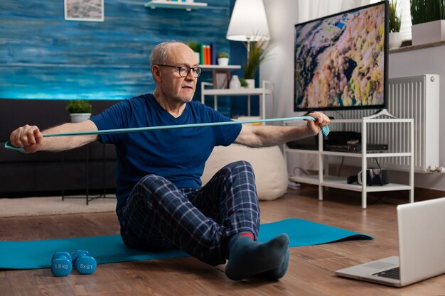 Pensioner working at body resistance exercising arms muscles using elastic band sitting on yoga mat