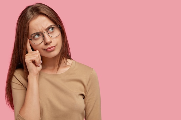 Foto gratuita una femmina adorabile pensierosa tiene il dito sulla tempia, essendo immersa nei pensieri, ha una faccia seria, ricorda qualcosa, posa contro il muro rosa