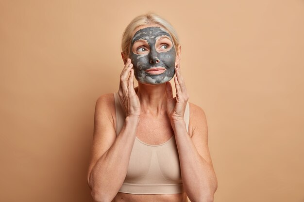 Pensive adorable middle aged lady has minimal makeup wears moisturising mask touches face gently dressed in casual top poses against beige wall