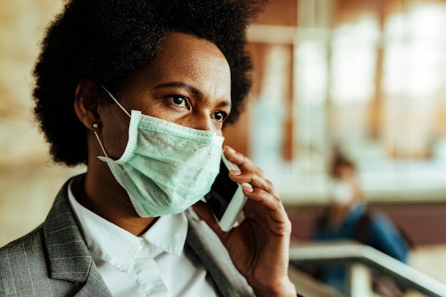 바이러스 전염병 동안 공공 기차역에서 전화 통화를 하는 동안 얼굴 마스크를 쓴 수심에 찬 아프리카계 미국인 여성 사업가