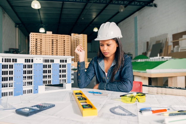 Foto gratuita donna afroamericana pensierosa nel casco di sicurezza vicino al modello di costruzione