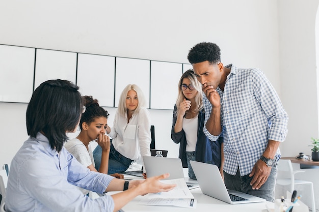 Pensive african office worker covering mouth with hand, while solving problem with computer. Team of asian and black web-programmers found a mistake in their project.