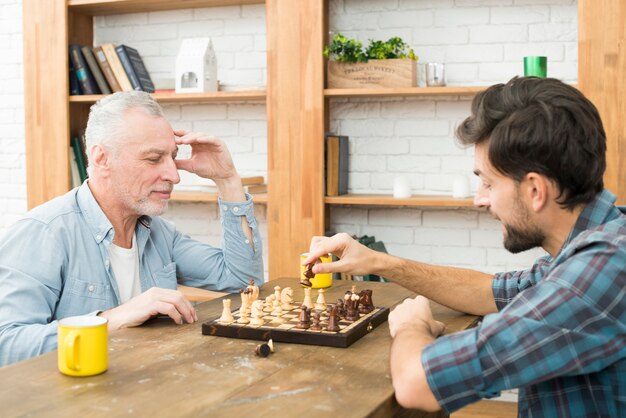 Pensive aged man and young guy playing chess at table in room