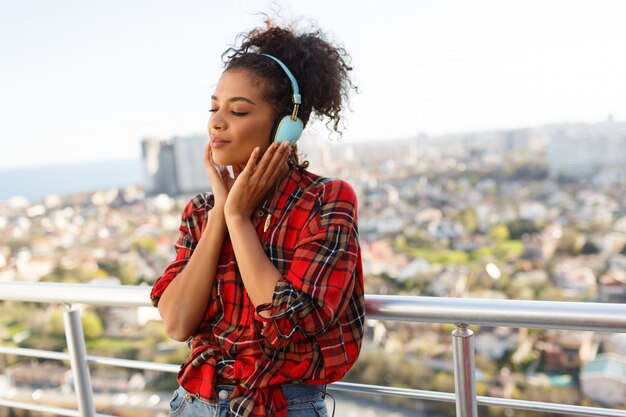 Pensive  American woman with dark skin  posing with earphones  on the rooftop. Urban landscape background.