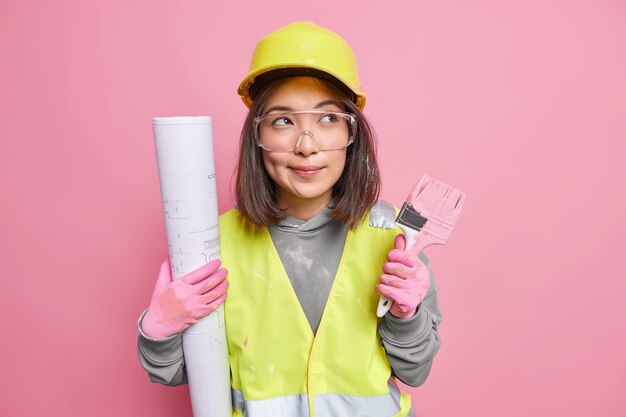 Pensive Asian female maintenance worker holds rolled blueprint painting brush looks away thoughtfully wears uniform poses on pink 