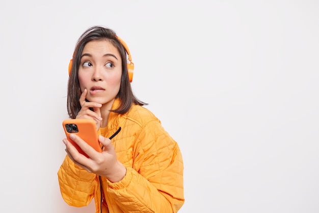 Pensive Asian female model bites lips has thoughtful look uses smartphone and stereo headphones for listening music dressed in jacket isolated over white wall with blank space on right